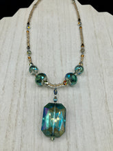 Load image into Gallery viewer, Make a bold statement with this shimmery aqua blue and gold tones glass beaded necklace. The large rectangular faceted shaped pendant creates a vivid contrast to the warm gold tones of the necklace, perfect for adding a rich pop of color to any Spring outfit. The draped design gives this piece an elegant, bohemian look. Measurements: Necklace is 20” in length,