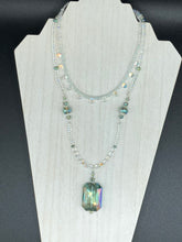 Load image into Gallery viewer, This stunning aqua blue and silver glass beaded necklace is a bold statement piece, composed of an adjustable necklace featuring blues, silver, and iridescent beads. Its shorter, collar style strand contains two-hole iridescent beads to keep its shape and adds a generous helping of iridescent teardrop glass beads for maximum sparkle. Its accompanying large, rectangular faceted pendant has a shimmering mix of gold, green, pink, and blue hues, and elegantly drapes across the neck.