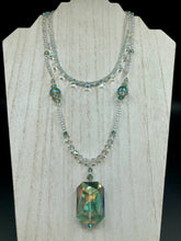 Load image into Gallery viewer, This stunning aqua blue and silver glass beaded necklace is a bold statement piece, composed of an adjustable necklace featuring blues, silver, and iridescent beads. Its shorter, collar style strand contains two-hole iridescent beads to keep its shape and adds a generous helping of iridescent teardrop glass beads for maximum sparkle. Its accompanying large, rectangular faceted pendant has a shimmering mix of gold, green, pink, and blue hues, and elegantly drapes across the neck.