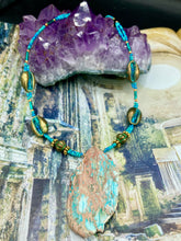 Load image into Gallery viewer, This stunning two-strand necklace features an array of glass beads in hues of green and blue turquoise, gold and green for a vibrant, colorful and bold look. The centerpiece is a large howlite stone chip dyed in turquoise color, accented by the bronze patina cowry beads. Measurements: Necklace is 17”