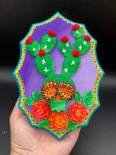 Load image into Gallery viewer, This handcrafted polymer clay wall art features a wood frame painted in a gradient from light to dark purple, creating a stunning sunset look. With a bright green skull cactus plant and bright red blooming flowers, this piece is sure to make a statement. Measurements: H 7" x W 5” x D 1 1/2"
