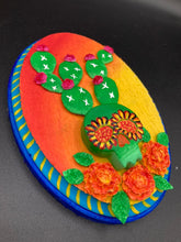 Load image into Gallery viewer, This handcrafted polymer clay wall art features a wood frame painted in a gradient from light yellow to dark red, creating a stunning sunset look. With a bright green skull cactus plant and bright pink blooming flowers. Boasting vivid marigold blooms and a deep royal blue frame to make a bold contrast with the warm background hue. this piece is sure to make a statement. Measurements: H 7" x W 5” x D 1 1/2"