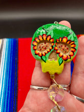 Load image into Gallery viewer, Nature inspired hand crafted polymer clay skull is painted in ombre colors from dark green to light yellow and decorated with flowers on back and over eyes. Light gold glass beaded chain with iridescent crystals. A perfect hanging Day of the Dead art piece to add to your ofrenda, hang next to your display of crystals or witchy altar, add to your skull collection or hang as an art piece on the wall or near a window. Measurements: H 1 1/2” x W 1 1/2” x D 3/4”