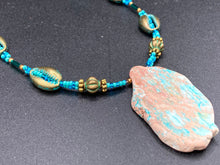 Load image into Gallery viewer, This stunning two-strand necklace features an array of glass beads in hues of green and blue turquoise, gold and green for a vibrant, colorful and bold look. The centerpiece is a large howlite stone chip dyed in turquoise color, accented by the bronze patina cowry beads. Measurements: Necklace is 17”