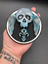 Load image into Gallery viewer, This handcrafted Dia De Los Muertos Calavera wall art decor is a unique way to bring life to your home and celebrates Mexican culture. Made with polymere clay and painted on a wooden frame, the beautiful blue skull is finished with wings and fleur de lis embellishments for a sophisticated look. Perfect for any room! Measurements: D 4"