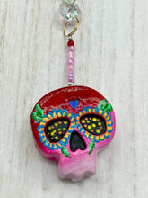 Load image into Gallery viewer, Nature inspired hand crafted polymer clay skull is painted in ombre colors from red, to light pink and decorated with flowers on back and over eyes. Heart rainbow hematite bead on forehead. Light pink glass beaded chain with iridescent crystals. A perfect hanging Day of the Dead art piece to add to your ofrenda, hang next to your display of crystals or witchy altar, add to your skull collection or hang as an art piece on the wall or near a window. Measurements: H 1 1/2” x W 1 1/2” x D 3/4”