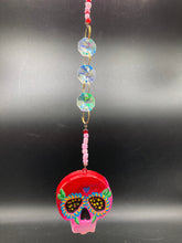 Load image into Gallery viewer, Nature inspired hand crafted polymer clay skull is painted in ombre colors from red, to light pink and decorated with flowers on back and over eyes. Heart rainbow hematite bead on forehead. Light pink glass beaded chain with iridescent crystals. A perfect hanging Day of the Dead art piece to add to your ofrenda, hang next to your display of crystals or witchy altar, add to your skull collection or hang as an art piece on the wall or near a window. Measurements: H 1 1/2” x W 1 1/2” x D 3/4”