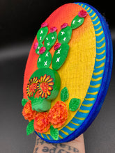 Load image into Gallery viewer, This handcrafted polymer clay wall art features a wood frame painted in a gradient from light yellow to dark red, creating a stunning sunset look. With a bright green skull cactus plant and bright pink blooming flowers. Boasting vivid marigold blooms and a deep royal blue frame to make a bold contrast with the warm background hue. this piece is sure to make a statement. Measurements: H 7" x W 5” x D 1 1/2"