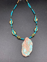 Load image into Gallery viewer, This stunning two-strand necklace features an array of glass beads in hues of green and blue turquoise, gold and green for a vibrant, colorful and bold look. The centerpiece is a large howlite stone chip dyed in turquoise color, accented by the bronze patina cowry beads. Measurements: Necklace is 17”