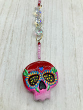 Load image into Gallery viewer, Nature inspired hand crafted polymer clay skull is painted in ombre colors from red, to light pink and decorated with flowers on back and over eyes. Heart rainbow hematite bead on forehead. Light pink glass beaded chain with iridescent crystals. A perfect hanging Day of the Dead art piece to add to your ofrenda, hang next to your display of crystals or witchy altar, add to your skull collection or hang as an art piece on the wall or near a window. Measurements: H 1 1/2” x W 1 1/2” x D 3/4”