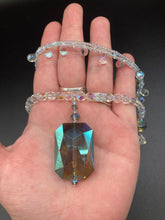 Load image into Gallery viewer, This stunning aqua blue and silver glass beaded necklace is a bold statement piece, composed of an adjustable necklace featuring blues, silver, and iridescent beads. Its shorter, collar style strand contains two-hole iridescent beads to keep its shape and adds a generous helping of iridescent teardrop glass beads for maximum sparkle. Its accompanying large, rectangular faceted pendant has a shimmering mix of gold, green, pink, and blue hues, and elegantly drapes across the neck.