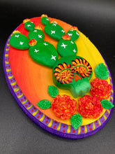 Load image into Gallery viewer, This handcrafted polymer clay wall art features a wood frame painted in a gradient from light yellow to dark red, creating a stunning sunset look. With a bright green skull cactus plant and bright yellow/red blooming flowers. Boasting vivid marigold blooms and a deep purple frame to make a bold contrast with the warm background hue. this piece is sure to make a statement. Measurements: H 7" x W 5” x D 1 1/2"