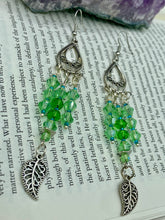 Load image into Gallery viewer, This large set of chandelier earrings features three long strands of shimmering glass beads in various light green and blue hues, accented with an intricate silver-plated leaf charm at the bottom for an added touch of movement. Versatile and perfect for whimsical, hippie-inspired, nature-loving looks. Measurements: 3 3/4” ” in length