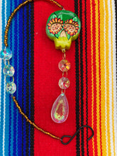 Load image into Gallery viewer, Nature inspired hand crafted polymer clay skull is painted in ombre colors from dark green to light yellow and decorated with flowers on back and over eyes. Light gold glass beaded chain with iridescent crystals. A perfect hanging Day of the Dead art piece to add to your ofrenda, hang next to your display of crystals or witchy altar, add to your skull collection or hang as an art piece on the wall or near a window. Measurements: H 1 1/2” x W 1 1/2” x D 3/4”