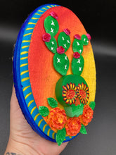 Load image into Gallery viewer, This handcrafted polymer clay wall art features a wood frame painted in a gradient from light yellow to dark red, creating a stunning sunset look. With a bright green skull cactus plant and bright pink blooming flowers. Boasting vivid marigold blooms and a deep royal blue frame to make a bold contrast with the warm background hue. this piece is sure to make a statement. Measurements: H 7" x W 5” x D 1 1/2"