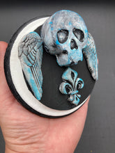 Load image into Gallery viewer, This handcrafted Dia De Los Muertos Calavera wall art decor is a unique way to bring life to your home and celebrates Mexican culture. Made with polymere clay and painted on a wooden frame, the beautiful blue skull is finished with wings and fleur de lis embellishments for a sophisticated look. Perfect for any room! Measurements: D 4"