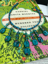 Load image into Gallery viewer, This necklace features vibrant teal quartzite and glass beads. The quartzite beads have a captivating swirl pattern, combining dark red and dark blue hues. It has an ethereal feel thanks to the various shades of blues, golds, and greens used to create a two-strand beaded design. For a dramatic finish, seven strands are added, completed with rectangular rainbow hematite beads for texture and depth. Both strands are held together with double-hole flat matte gold beads to keep a collar shape.