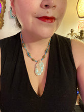 Load image into Gallery viewer, This stunning two-strand necklace features an array of glass beads in hues of green and blue turquoise, gold and green for a vibrant, colorful and bold look. The centerpiece is a large howlite stone chip dyed in turquoise color, accented by the bronze patina cowry beads. Measurements: Necklace is 17”
