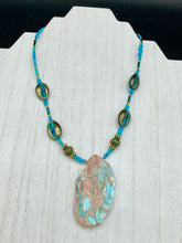 Load image into Gallery viewer, This stunning two-strand necklace features an array of glass beads in hues of green and blue turquoise, gold and green for a vibrant, colorful and bold look. The centerpiece is a large howlite stone chip dyed in turquoise color, accented by the bronze patina cowry beads. Measurements: Necklace is 17”