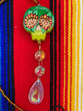 Load image into Gallery viewer, Nature inspired hand crafted polymer clay skull is painted in ombre colors from dark green to light yellow and decorated with flowers on back and over eyes. Light gold glass beaded chain with iridescent crystals. A perfect hanging Day of the Dead art piece to add to your ofrenda, hang next to your display of crystals or witchy altar, add to your skull collection or hang as an art piece on the wall or near a window. Measurements: H 1 1/2” x W 1 1/2” x D 3/4”