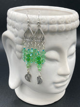 Load image into Gallery viewer, This large set of chandelier earrings features three long strands of shimmering glass beads in various light green and blue hues, accented with an intricate silver-plated leaf charm at the bottom for an added touch of movement. Versatile and perfect for whimsical, hippie-inspired, nature-loving looks. Measurements: 3 3/4” ” in length