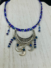 Load image into Gallery viewer, This stylish choker necklace showcases a large half moon pendant, adorned with glass beads and small, dangling crescent moon charms. Its eye-catching hues, of blue and silver, are complemented with delicate tints of purple and pink. Boasting an accentuated look and ample motion, this piece is sure to impress. Measurements: Necklace is 16 1/2”adjustable up to 18"
