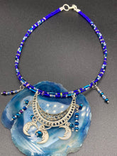 Load image into Gallery viewer, This stylish choker necklace showcases a large half moon pendant, adorned with glass beads and small, dangling crescent moon charms. Its eye-catching hues, of blue and silver, are complemented with delicate tints of purple and pink. Boasting an accentuated look and ample motion, this piece is sure to impress. Measurements: Necklace is 16 1/2”adjustable up to 18"