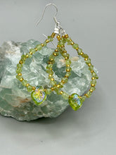 Load image into Gallery viewer, These natural peridot chips earrings feature a gold glass beading on their hoops, creating an eye-catching light green tone. A pressed glass leaf charm is also included, boasting an iridescent shimmer finish with subtle hints of gold, purple, blue, and pink. Measurements: 2 1/2" length