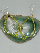 Load image into Gallery viewer, These natural peridot chips earrings feature a gold glass beading on their hoops, creating an eye-catching light green tone. A pressed glass leaf charm is also included, boasting an iridescent shimmer finish with subtle hints of gold, purple, blue, and pink. Measurements: 2 1/2" length