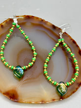 Load image into Gallery viewer, This pair of earrings features a dark green, pressed glass leaf charm in an iridescent finish that gives off subtle hues of gold, purple, blue, and pink. Complemented with green and gold glass beaded hoop earrings, this set is sure to sparkle. Measurements: 2 3/4" length