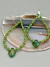 Load image into Gallery viewer, This pair of earrings features a dark green, pressed glass leaf charm in an iridescent finish that gives off subtle hues of gold, purple, blue, and pink. Complemented with green and gold glass beaded hoop earrings, this set is sure to sparkle. Measurements: 2 3/4" length