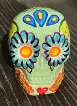 Load image into Gallery viewer, Inspired by nature, this ceramic skull statuette is full of vibrant color and intricate details. The primary color is soft, light periwinkle blue with Multi-color floral motifs that are painted across the skull, along with hearts and leaves and lively colorful accents around the entire skull.