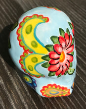 Load image into Gallery viewer, This ceramic skull statuette is a stunning piece of art inspired by nature. Its soft periwinkle blue hue is decorated with multi-color floral motifs, hearts, leaves and colorful accents, creating a beautiful piece of hand-painted, Day of the Dead-themed art. H 2 3/4” x W 2” x D 2”