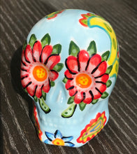 Load image into Gallery viewer, Inspired by nature, this ceramic skull statuette is full of vibrant color and intricate details. The primary color is soft, light periwinkle blue with Multi-color floral motifs that are painted across the skull, along with hearts and leaves and lively colorful accents around the entire skull.