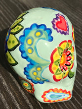 Load image into Gallery viewer, Inspired by nature, this ceramic skull statuette is full of vibrant color and intricate details. The primary color is soft pastel green with Multi-color floral motifs that are painted across the skull, along with hearts and leaves and lively colorful accents around the entire skull.