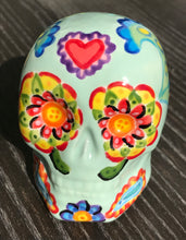 Load image into Gallery viewer, Inspired by nature, this ceramic skull statuette is full of vibrant color and intricate details. The primary color is soft pastel green with Multi-color floral motifs that are painted across the skull, along with hearts and leaves and lively colorful accents around the entire skull.
