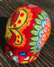 Load image into Gallery viewer, This ceramic skull statuette is a stunning piece of art inspired by nature. Its vibrant red hue is decorated with multi-color floral motifs, hearts, leaves and colorful accents, creating a beautiful piece of hand-painted, Day of the Dead-themed art. Measurements: H 2 3/4” x W 2” x D 2”