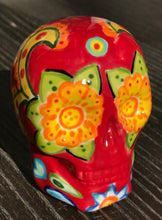 Load image into Gallery viewer, This ceramic skull statuette is a stunning piece of art inspired by nature. Its vibrant red hue is decorated with multi-color floral motifs, hearts, leaves and colorful accents, creating a beautiful piece of hand-painted, Day of the Dead-themed art. Measurements: H 2 3/4” x W 2” x D 2”