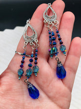 Load image into Gallery viewer, This set of large, eye-catching chandelier-style earrings draw inspiration from the vibrant array of colors found in the galaxy and cosmos. Featuring three strands of shimmering glass beads in bold shades of blue, purple, gold, and green, it's an essential piece for any jewelry collection. Measurements: 3 1/2” in length