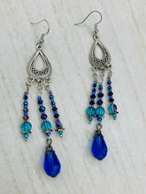 Load image into Gallery viewer, This set of large, eye-catching chandelier-style earrings draw inspiration from the vibrant array of colors found in the galaxy and cosmos. Featuring three strands of shimmering glass beads in bold shades of blue, purple, gold, and green, it's an essential piece for any jewelry collection. Measurements: 3 1/2” in length