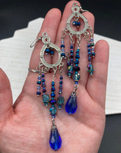 Load image into Gallery viewer, This beautifully large and bold set of chandelier earrings were inspired by the vibrant and beautiful galaxy and cosmos full of bright and stunning hues of blues and purples. I created this set of earrings with strands of glass beads full of shimmer and bold hues of blues, purples and a tint of gold and greens colors. This is a long, large, and bold piece of jewelry have a lot of shimmer and movement, a “must have” to your collection. Measurements: 4” in length