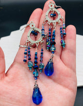 Load image into Gallery viewer, This beautifully large and bold set of chandelier earrings were inspired by the vibrant and beautiful galaxy and cosmos full of bright and stunning hues of blues and purples. I created this set of earrings with strands of glass beads full of shimmer and bold hues of blues, purples and a tint of gold and greens colors. This is a long, large, and bold piece of jewelry have a lot of shimmer and movement, a “must have” to your collection. Measurements: 4” in length