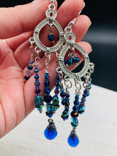 Load image into Gallery viewer, This beautifully large and bold set of chandelier earrings were inspired by the vibrant and beautiful galaxy and cosmos full of bright and stunning hues of blues and purples. I created this set of earrings with strands of glass beads full of shimmer and bold hues of blues, purples and a tint of gold and greens colors. This is a long, large, and bold piece of jewelry have a lot of shimmer and movement, a “must have” to your collection. Measurements: 4” in length