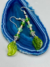 Load image into Gallery viewer, Long strand of glittering light green and gold glass beads, as well as a leaf-shaped glass charm at the bottom, combine to create a highly dynamic effect. These versatile earrings, perfect for a whimsical fairy princess, good witch, or nature-loving hippie look, draw on nature-inspired designs, providing the ideal finishing touch to any ensemble. Measurements: 2 1/4” in length