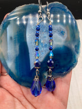 Load image into Gallery viewer, This stunning, one of a kind pair of celestial beaded earrings is a bold addition to any jewelry collection. Crafted with vibrant blue glass beads, the color contains varied shades of blue, purple, and hints of gold to create unique texture. The design is inspired by the awe-inspiring beauty of the majestic galaxy. Measurements: 2 3/4 ” in length