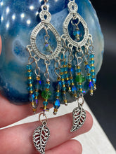Load image into Gallery viewer, These large chandelier-style earrings feature blues, greens, and iridescent glass beads as well as a finely crafted silver plated leaf charm for added detail. Perfect for bohemian-inspired looks, these versatile earrings can elevate any wardrobe from whimsical hippie to boho chic. Measurements: 3 1/4” in length