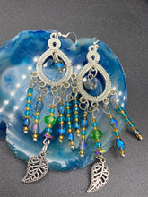 Load image into Gallery viewer, These large chandelier-style earrings feature blues, greens, and iridescent glass beads as well as a finely crafted silver plated leaf charm for added detail. Perfect for bohemian-inspired looks, these versatile earrings can elevate any wardrobe from whimsical hippie to boho chic. Measurements: 3 1/4” in length