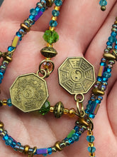 Load image into Gallery viewer, This collar-style necklace is a showstopper, beaded with stylish glass beads in green and blue tones, featuring contrasting yin-yang charms with differing sides. Rectangular rainbow hematite beads add complexity and depth, and double-hole matte gold beads keep the collar shape complete. It gracefully drapes around the neck, making a timeless statement of elegance.  Measurements:  Necklace is 14”, can be adjusted up to 18"