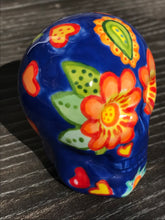 Load image into Gallery viewer, Inspired by nature, this ceramic skull statuette is full of vibrant color and intricate details. The primary color is royal blue with multi-color floral motifs that are painted across the skull, along with hearts and leaves and lively colorful accents around the entire skull.