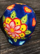 Load image into Gallery viewer, Inspired by nature, this ceramic skull statuette is full of vibrant color and intricate details. The primary color is royal blue with multi-color floral motifs that are painted across the skull, along with hearts and leaves and lively colorful accents around the entire skull.